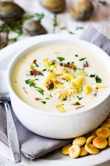 A large white soup bowl filled with New England Clam Chowder on a plate with oyster crackers and spoon next to it.