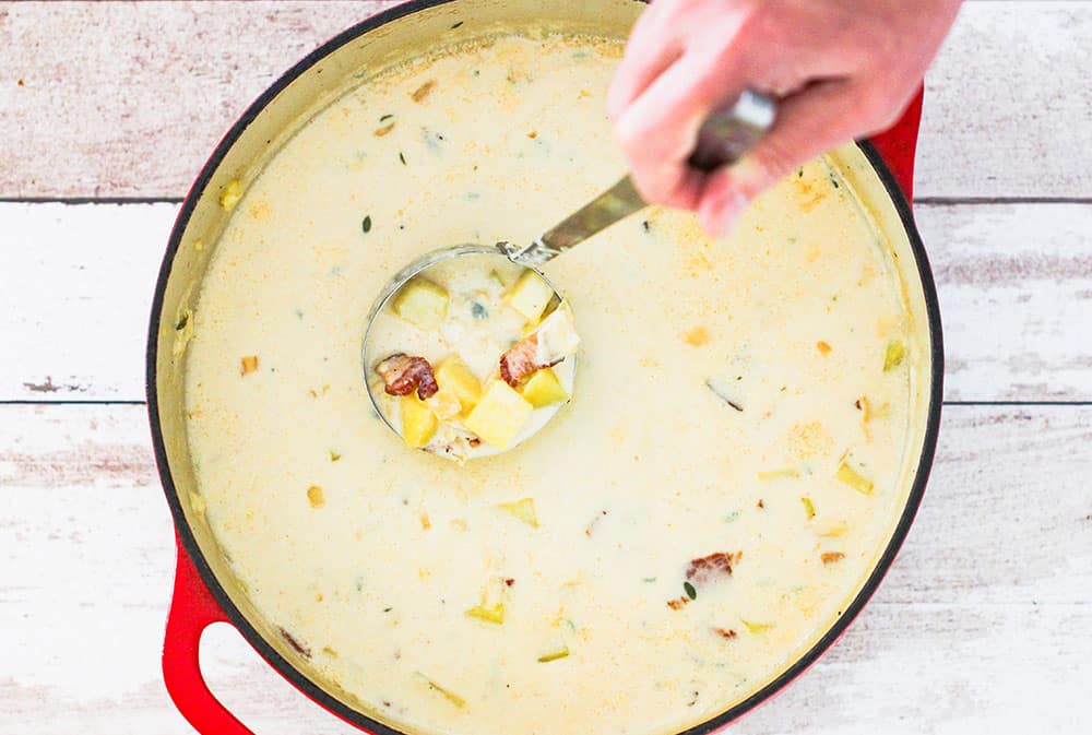 An overhead view of a large pot of New England Clam Chowder with a hand holding a ladle in the bowl. 