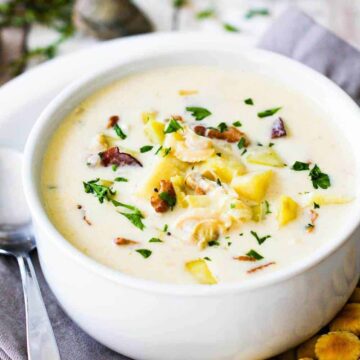 A large white soup bowl filled with New England Clam Chowder on a plate with oyster crackers and spoon next to it.