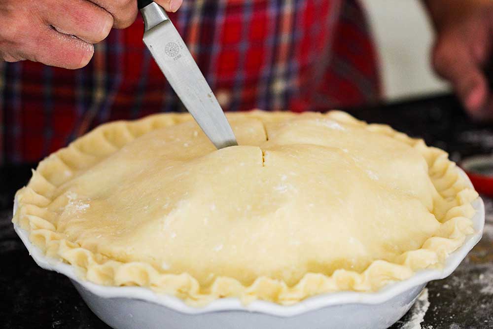 A hand using a knife to cut slits in the top of an uncooked homemade apple pie.