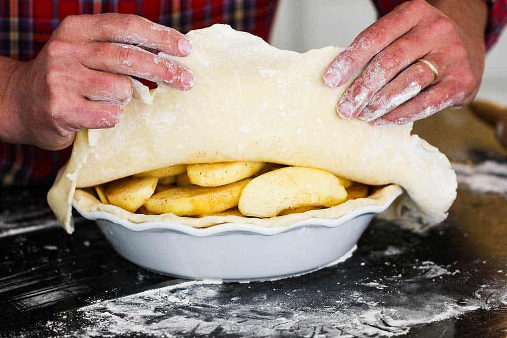 Two hands adding a top layer of pie dough over a pie dish filled with apples.