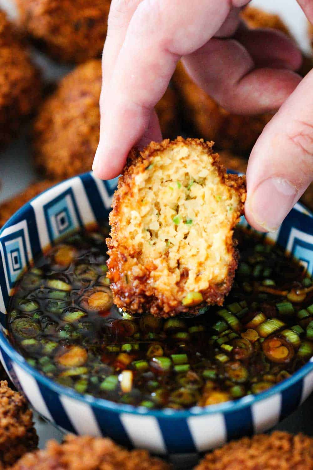 A hand dipping a partial fried shrimp ball into a bowl of spicy dipping sauce in a colorful bowl.