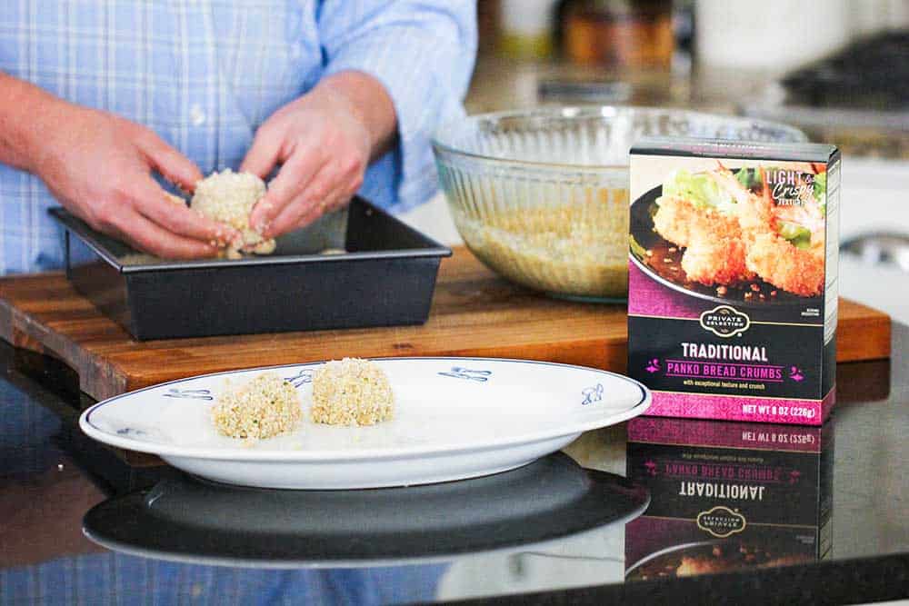 Two hands forming batter into balls and rolling in bread crumbs in a pan with a dish next to it.