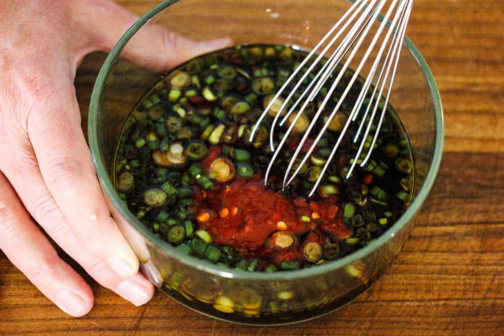 A hand holding a glass bowl of spicy Asian dipping sauce with a whisk in it.