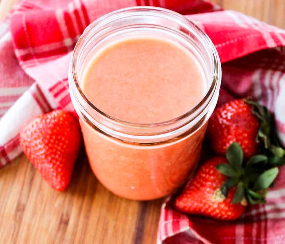 A jar of strawberry vinaigrette next to three fresh strawberries and a red napkin.