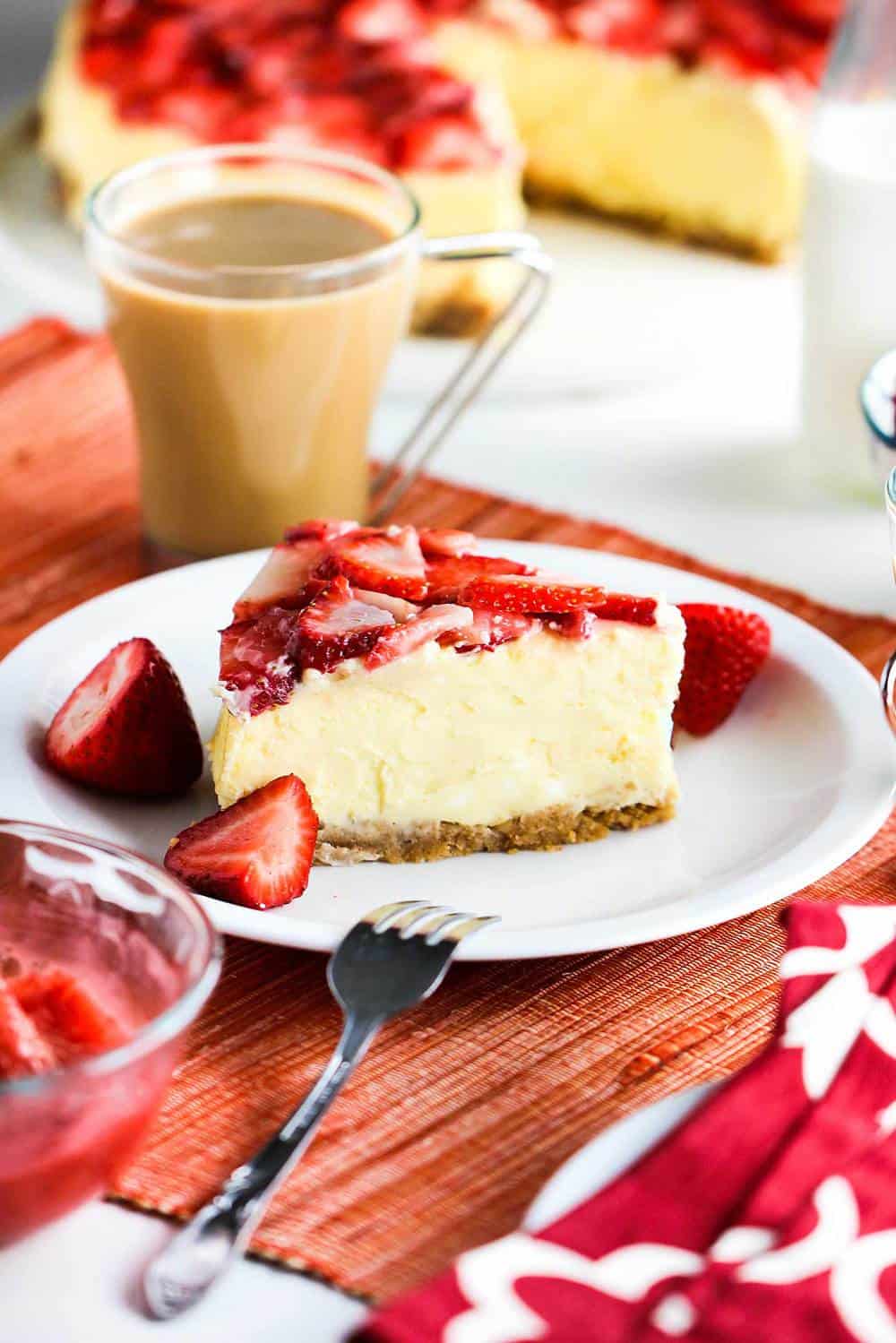 A slice of New York Cheese Cake with Strawberry Glaze sitting next to a cup of coffee and fresh strawberries.