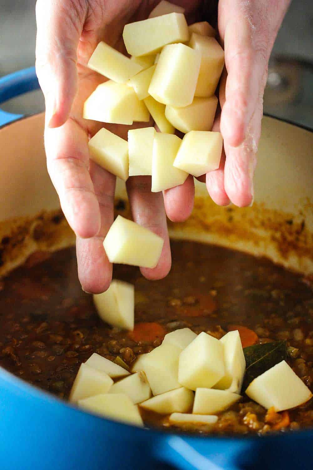 Two hands dropping cubed potatoes into a Dutch oven filled with lentil soup.
