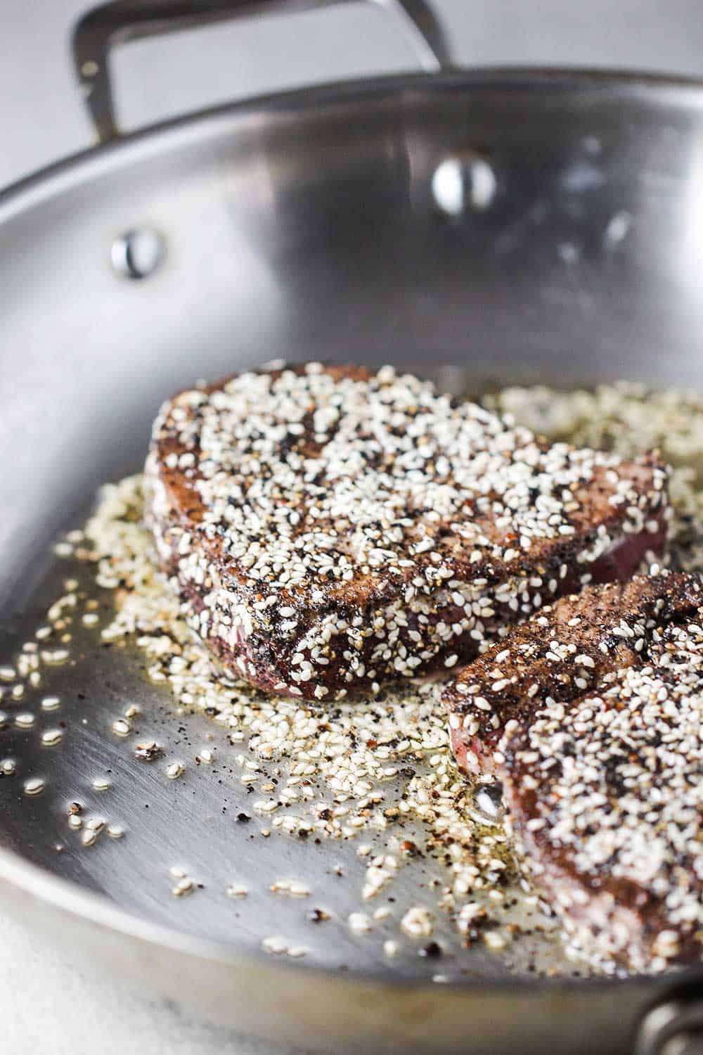 Two tuna steaks covered in black pepper and sesame seeds being seared in a large sliver skillet.