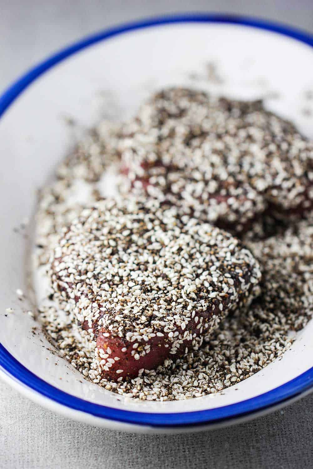 Two raw tuna steaks dredged in ground peppercorns and white sesame seeds on a platter.