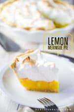 A single slice of a lemon meringue pie on a white plate with the whole pie in the background.