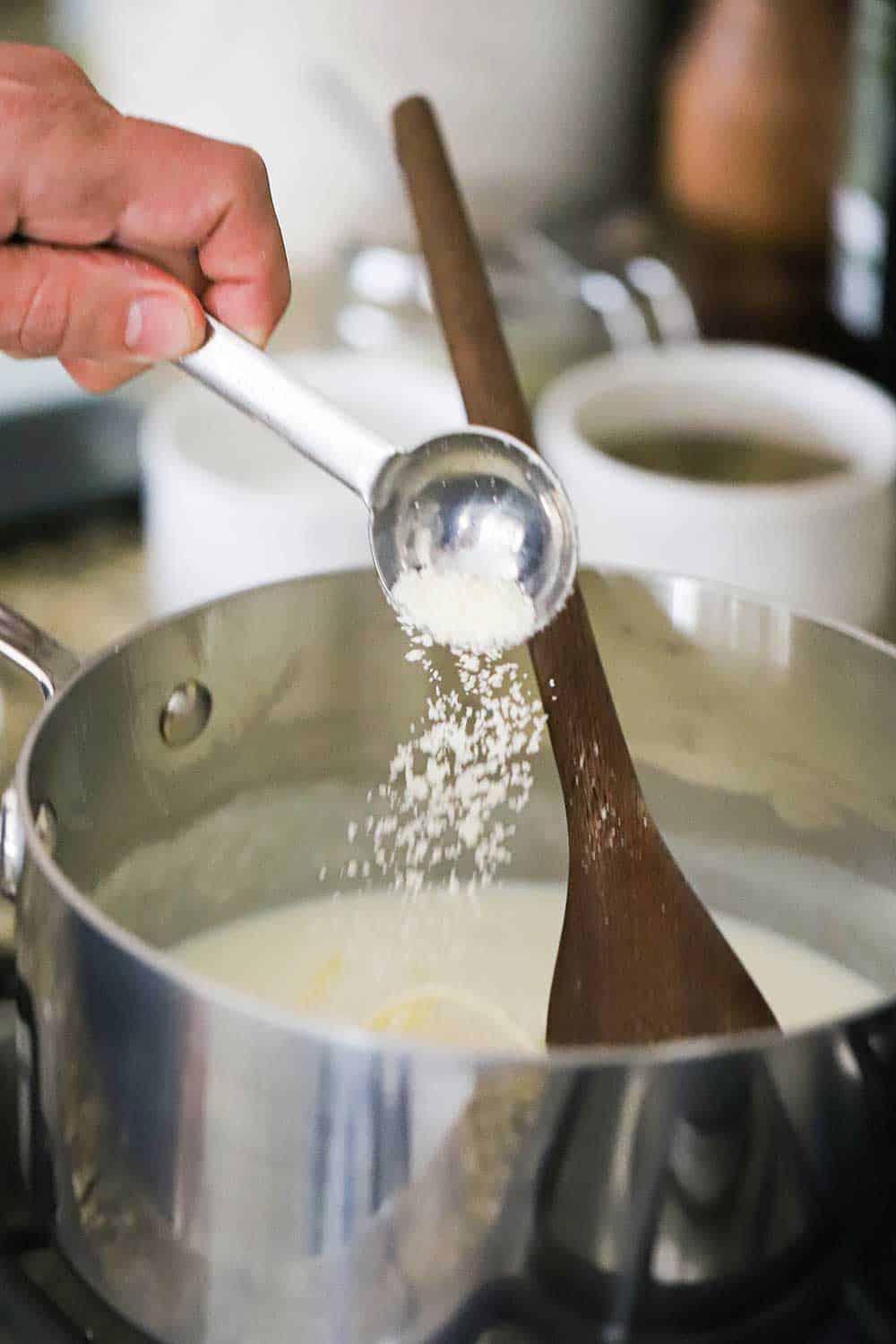 A person sprinkling grated Parmesan cheese from a tablespoon into a saucepan filled with Mornay sauce.