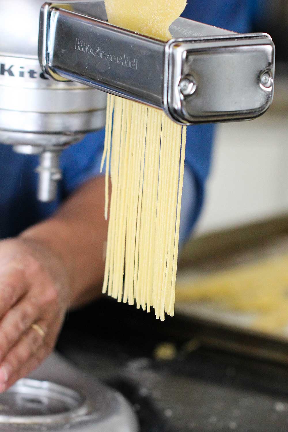 Pasta dough being cut by a pasta attachment on the side of a stand mixer.