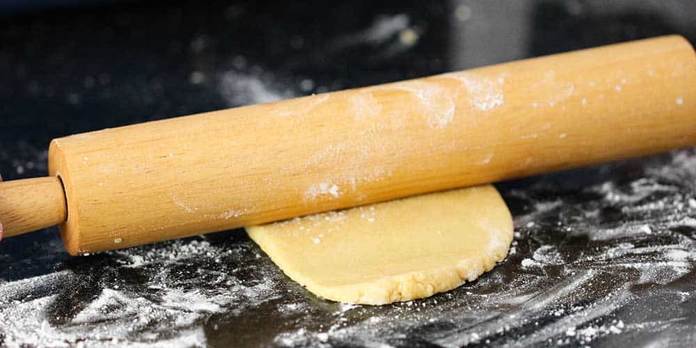 A rolling pin rolling out a piece of homemade pasta dough on a floured workspace.
