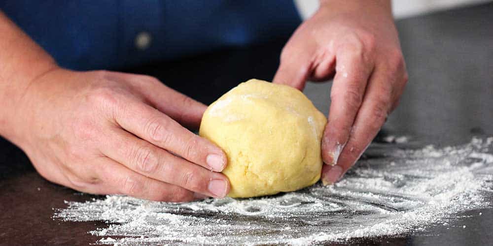 Two hands form pasta dough into a ball on a floured work surface.