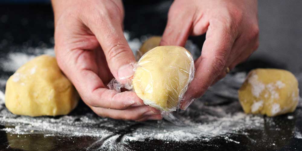 Two hands wrapping a piece of homemade pasta dough in plastic wrap.
