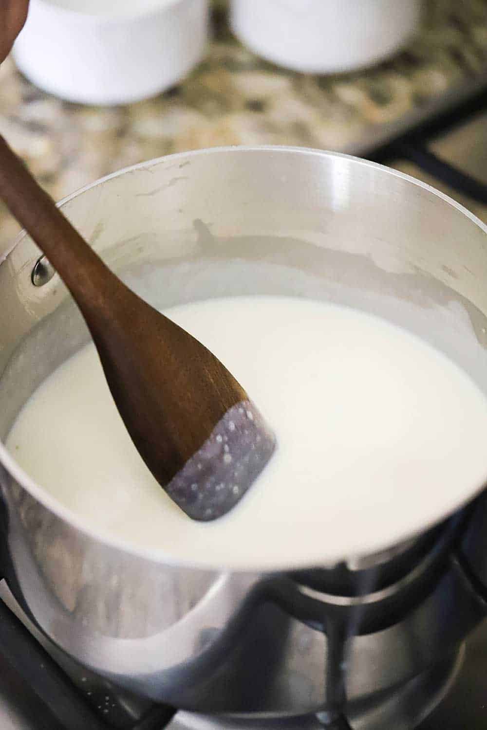 A stainless steel saucepan filled with a white roux being stirred with a flat wooden spoon.