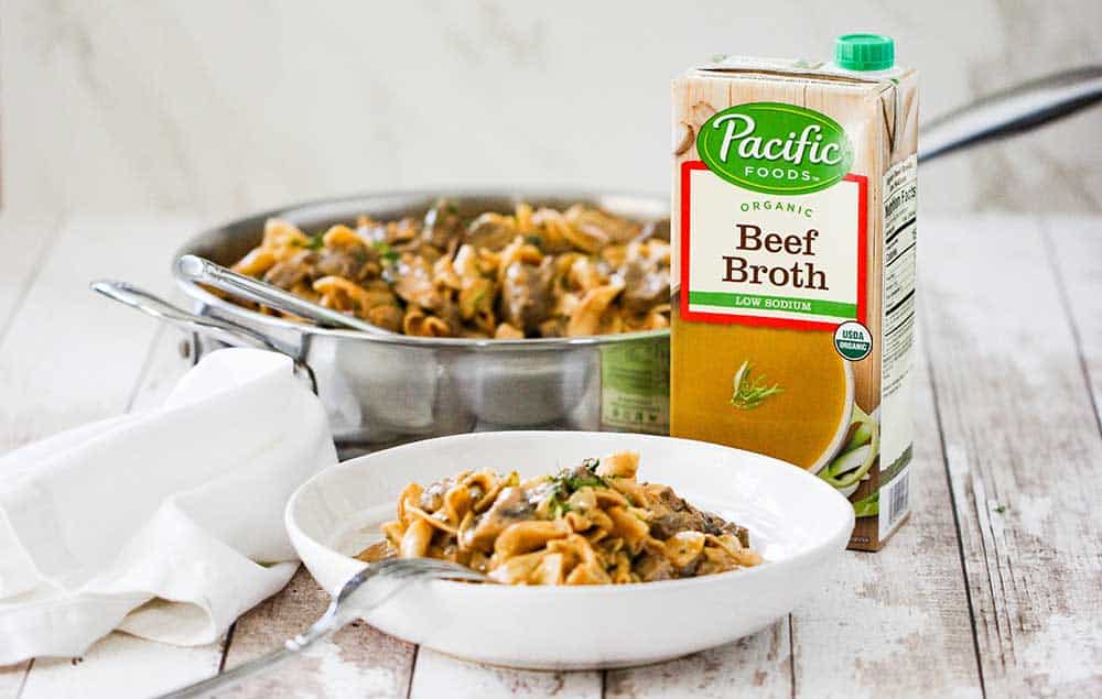 A white bowl of gourmet beef stroganoff with a box of beef broth in the background.
