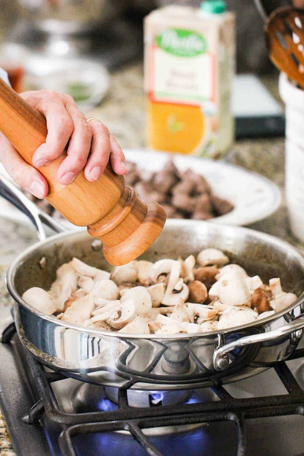 A pepper grinder being twisted into a pan of mushrooms on a stove.