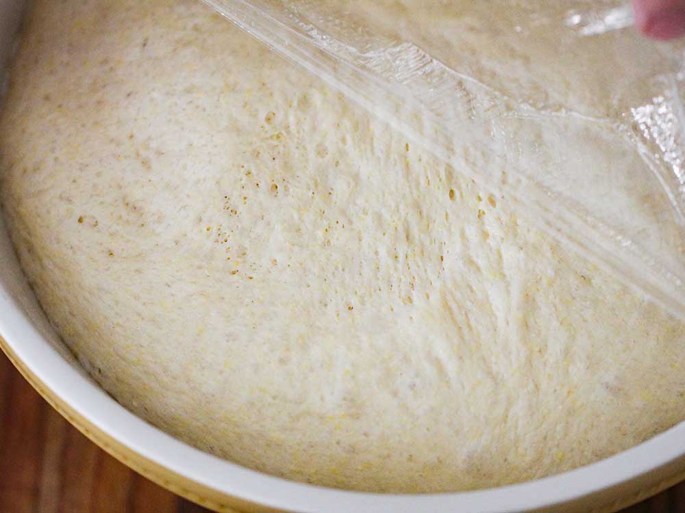 Risen pizza dough in a ceramic bowl with a hand pulling back plastic wrap on top.