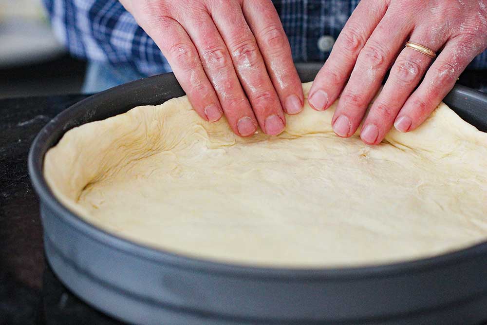 Two hands pressing pizza dough into a deep dish pizza pan.