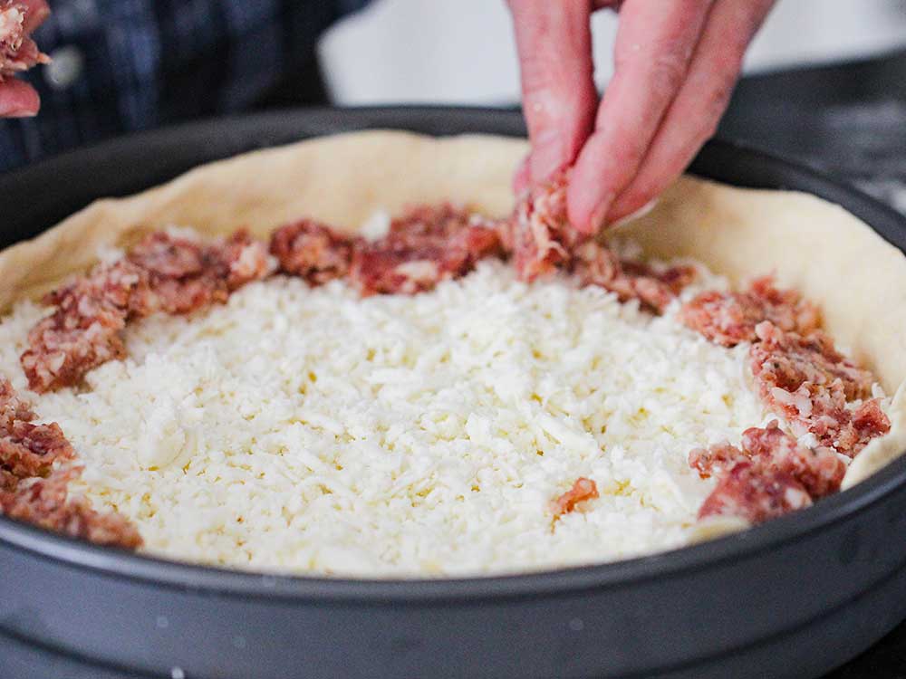 A hand layer pieces of Italian sausage over a bed of cheese in a pizza pan.