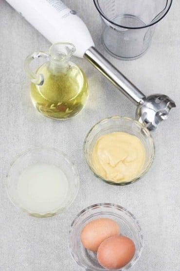 An overhead view of an immersion blender next to vessels filled with safflower oil, Dijon mustard, white wine vinegar, and 2 whole eggs.