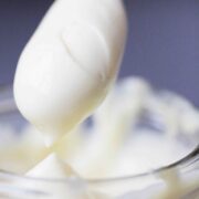 A spoon lifted from a clear container completely covered with homemade mayonnaise.