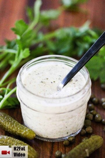 A small jar of homemade tartar sauce with a spoon it and pickles and capers nearby.