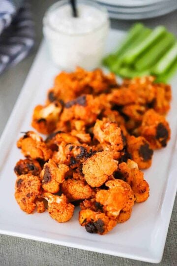 A rectangular white serving platter filled with buffalo cauliflower bites sitting next to a bowl of blue cheese dressing.