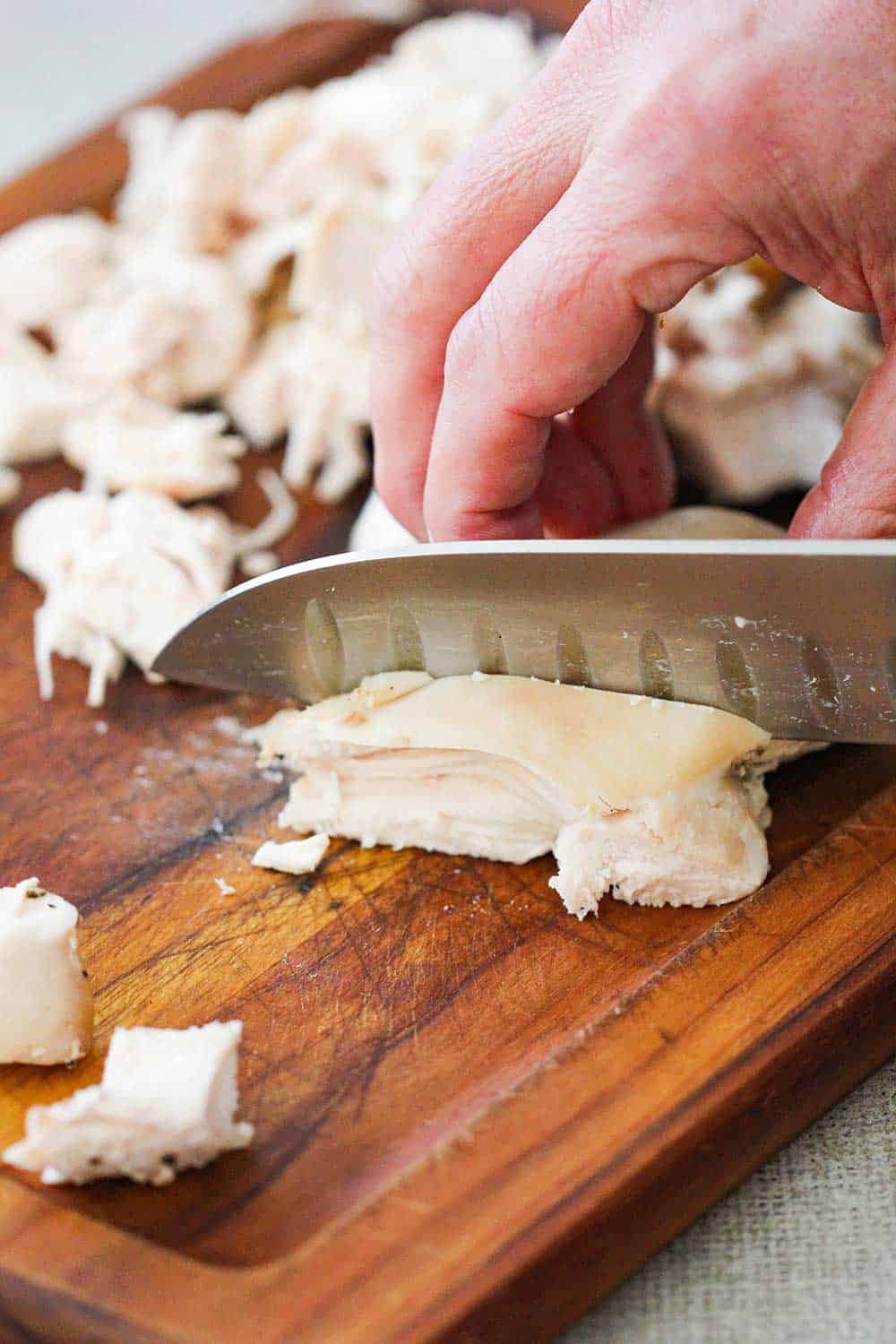 A hand using a large chef's knife to cube cooked chicken on a cutting board.