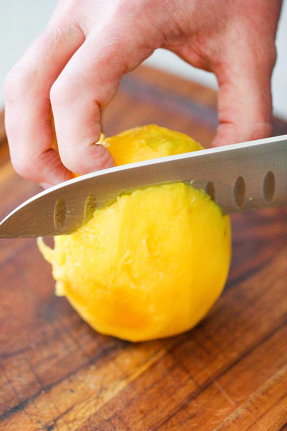 A hand using a large chef's knife to slice a fresh mango on a cutting board.