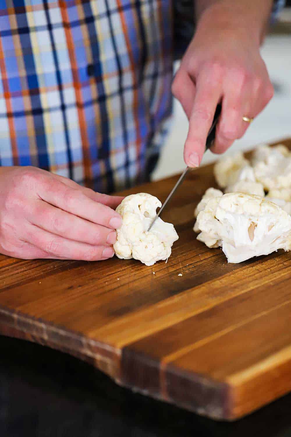 A person using a knife to cut a cauliflower heat into florets on a wooden cutting board.