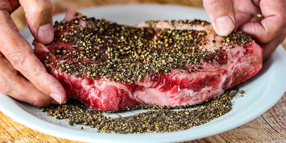 Spread the uncooked strip steaks in the cracked black peppercorns.
