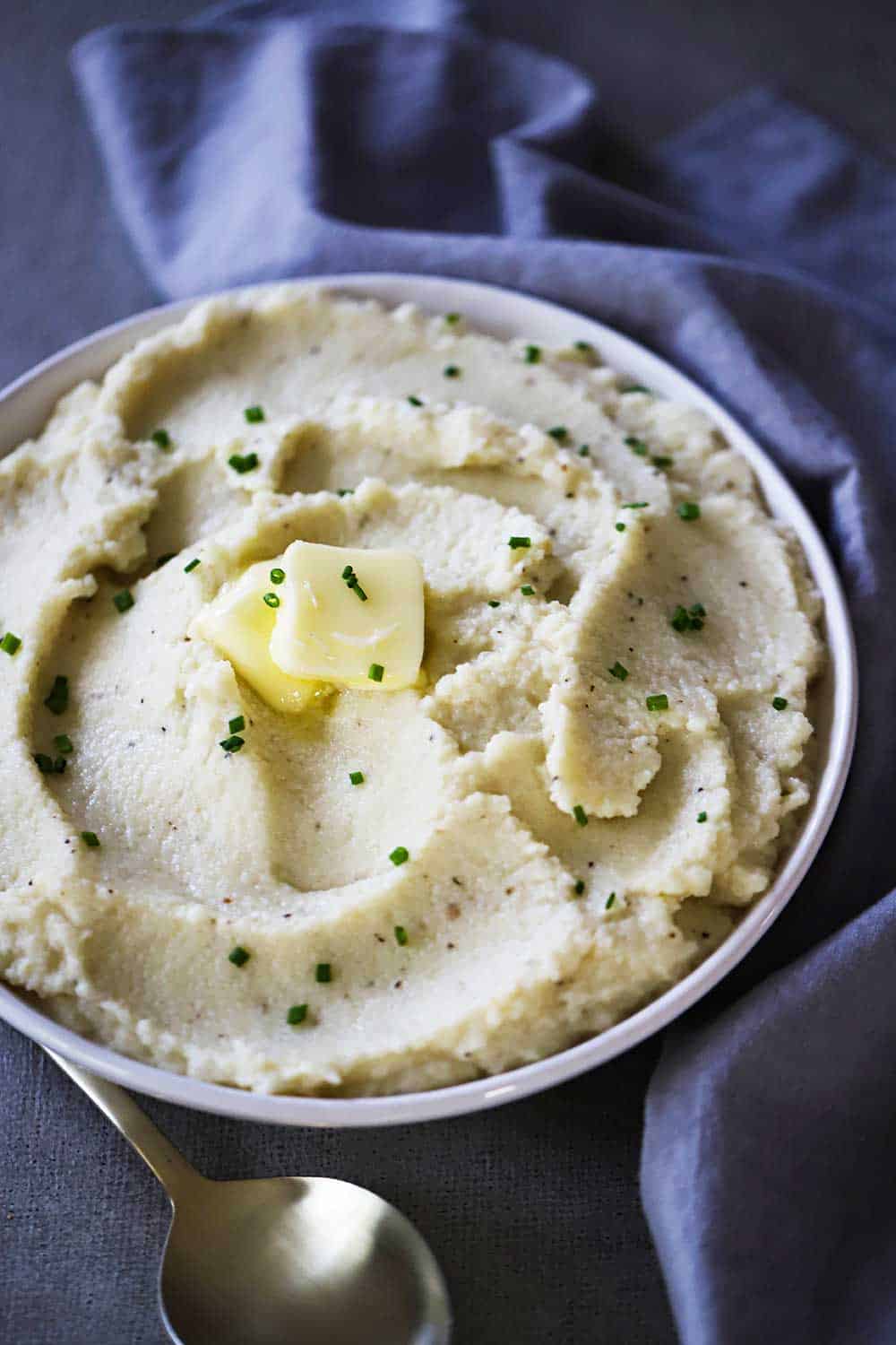 A white bowl filled with mashed cauliflower and roasted garlic with a pad of butter melting on top, all sitting next to a blue napkin and gold spoon.