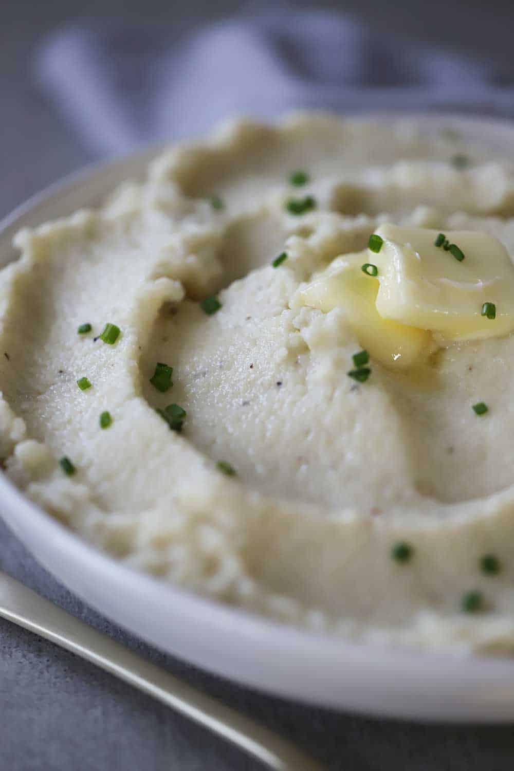 A circular white bowl filled with mashed cauliflower with roasted garlic topped with a pad of melting butter and snipped chives.