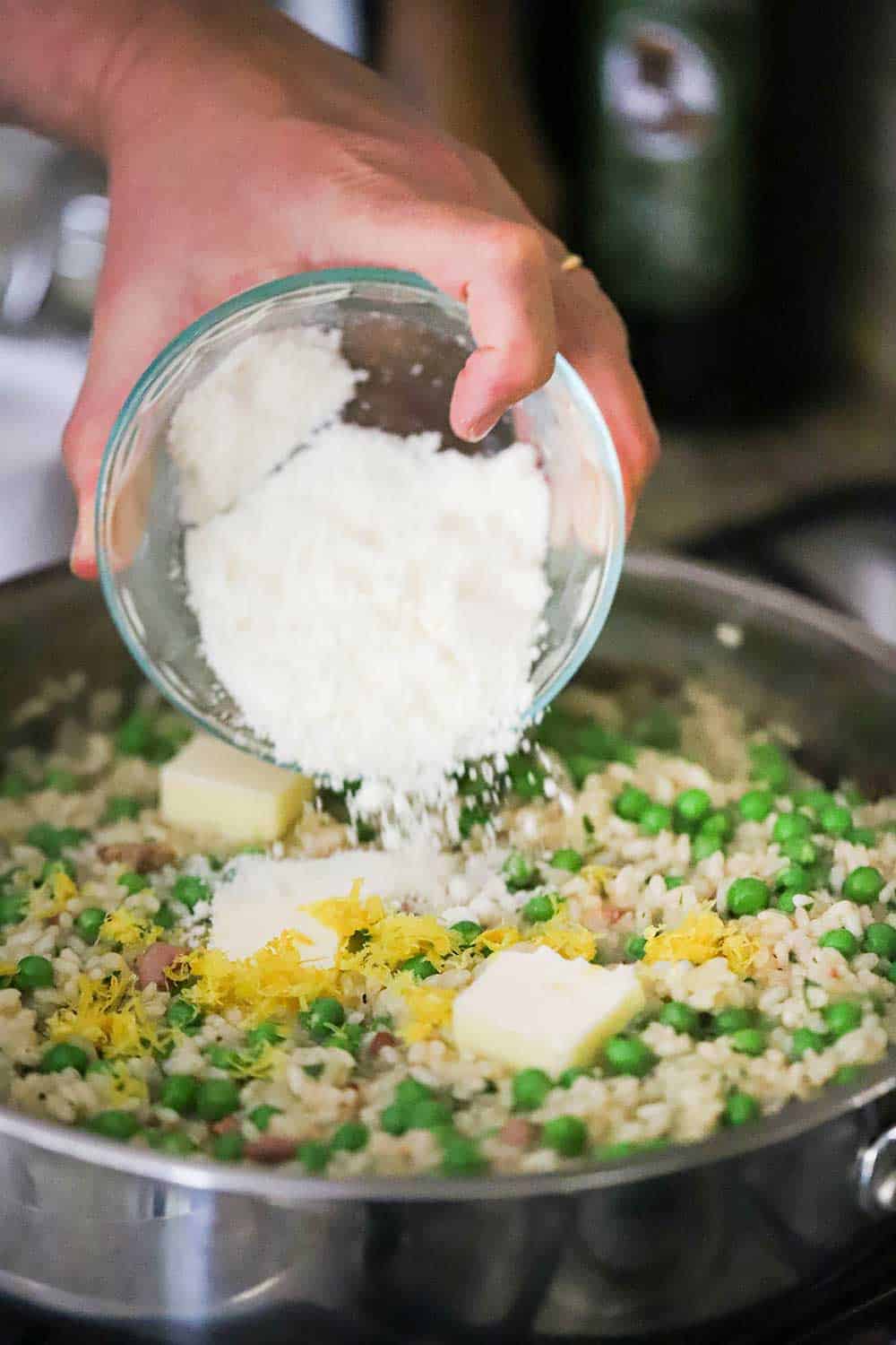 A person using their hand to transfer grated Parmesan cheese from a small bowl into a skillet of cooked risi e bisi.