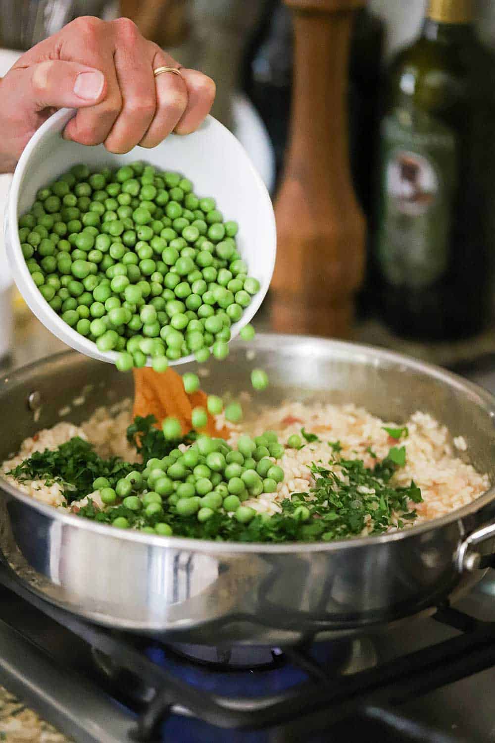 A person transferring shelled English peas from a small white bowl into a large skillet filled with cooked arborio rice.