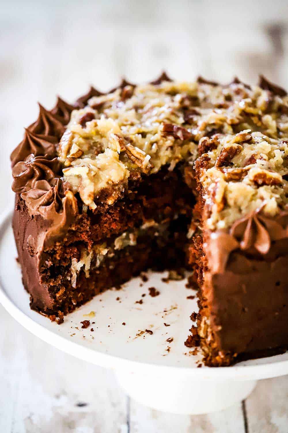 A German chocolate cake sitting on a white cake stand with a slice cut out of it.