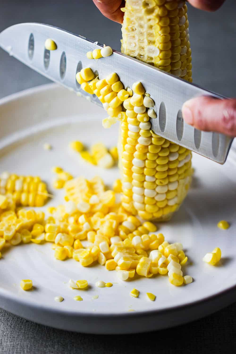 A hand using a large knife to remove corn kernels from an ear of corn.