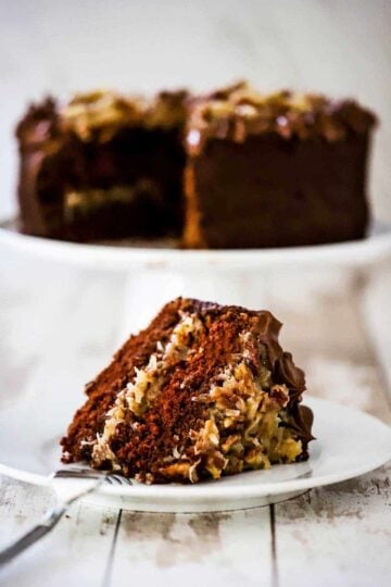 A slice of German chocolate cake on a white dessert plate in front of a cake stand with the cake on it.