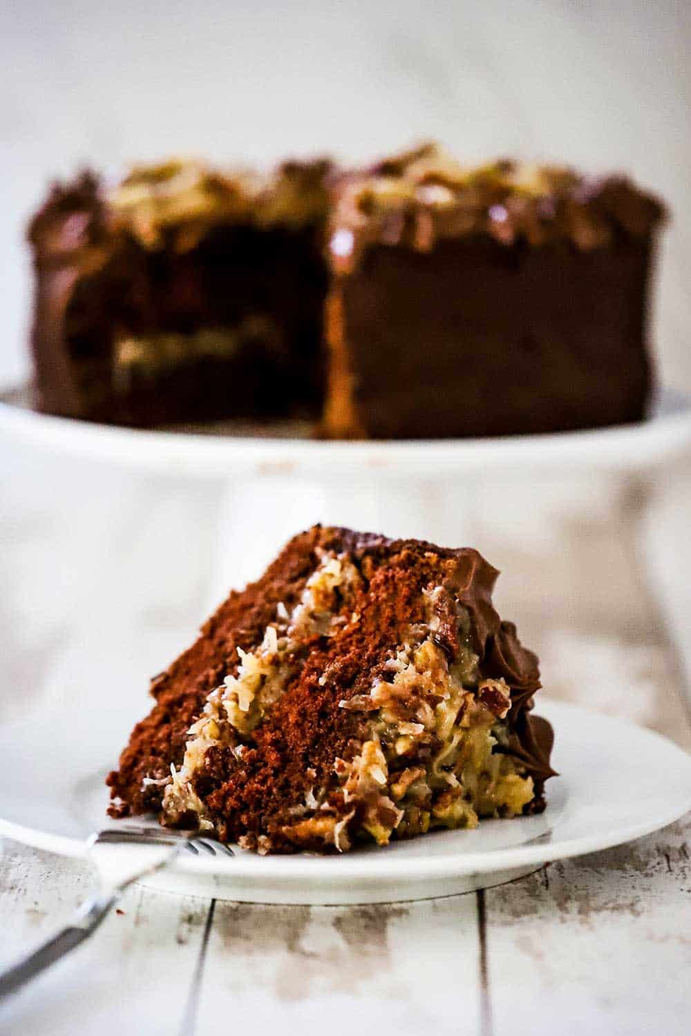 A slice of German chocolate cake on a white dessert plate in front of a cake stand with the cake on it.