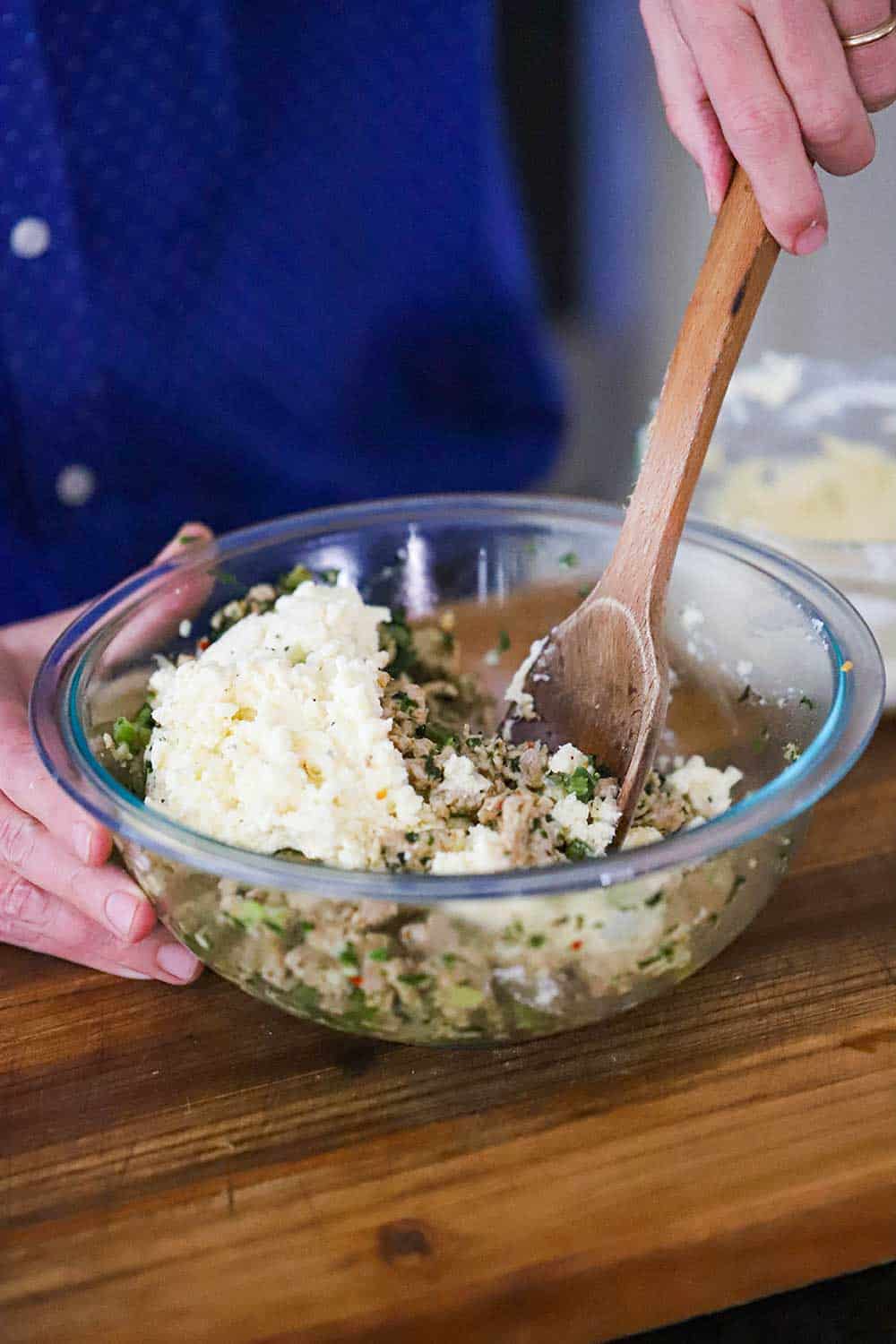 A person using a large wooden spoon to stir together a cooked chicken mixture in a glass bowl with a ricotta and mozzarella mixture.
