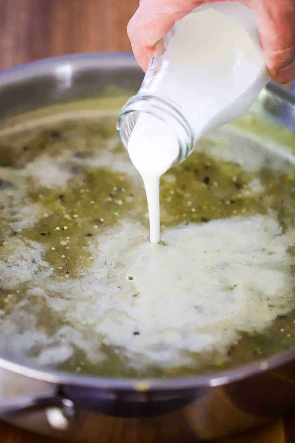 A hand pouring Mexican crema from a bottle into a skillet filled with hot verdi sauce.