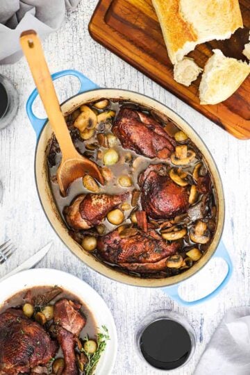 An overhead view of a large oval-shaped Dutch oven filled with Coq au Vin with a wooden spoon in and next to a cutting board topped with bread and next to a glass of red wine.