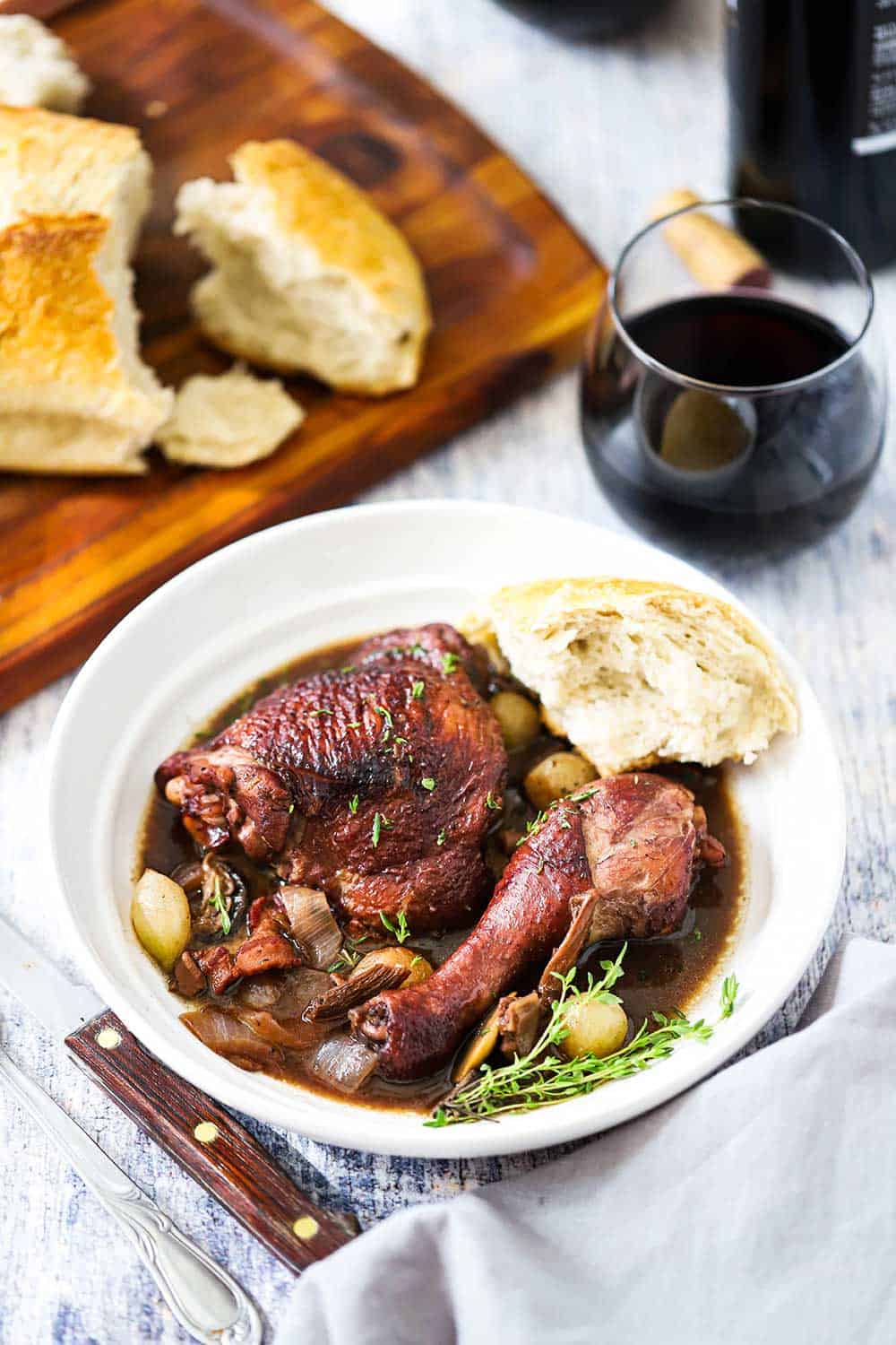 A white plate holding Coq au Vin with a piece of a loaf of bread next to it, and a stemless glass of red wine and a loaf of bread nearby.