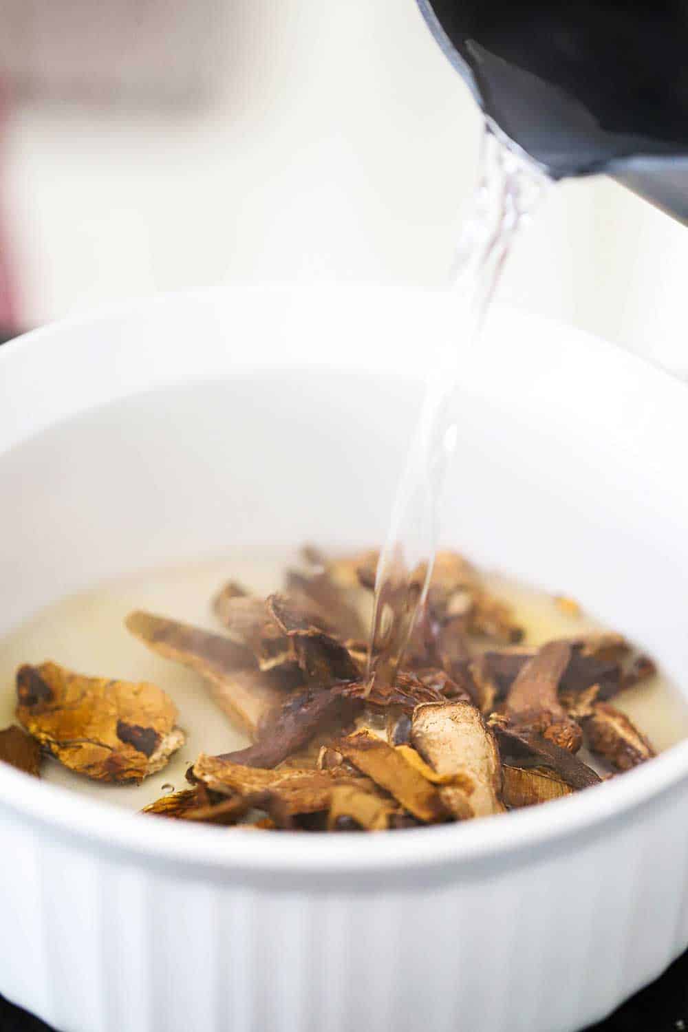 A circular white baking dish filled with dried porcini mushrooms with hot water being poured over them from a pot.
