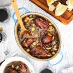 An overhead view of a large oval-shaped Dutch oven filled with Coq au Vin with a wooden spoon in and next to a cutting board topped with bread and next to a glass of red wine.