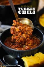 A helping of turkey chili being ladled into a dark blue bowl next to a piece of cornbread.