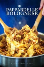 Two hands holding two wooden spoons tossing pappardelle bolognese in a large silver skillet.