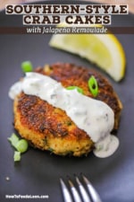 A close-up view of a Southern-style crab cake with a drizzle of jalepeño remoulade poured over the top and a lemon wedge next to it.